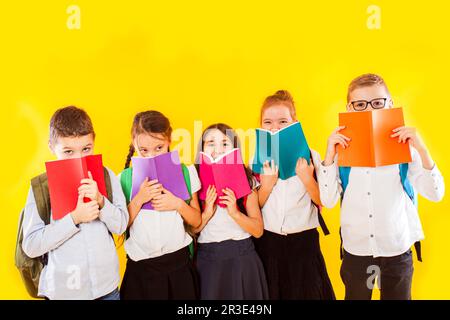 Fröhliche Schulkinder stehen mit bunten Büchern isoliert auf gelbem Hintergrund. Kinder verstecken sich hinter Büchern Stockfoto