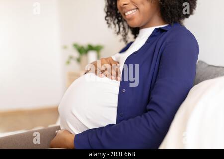 Glückliche schwarze Millennials-Frau in Haushaltskleidung mit großem Bauch genießt die Schwangerschaft, sitzt auf dem Bett Stockfoto
