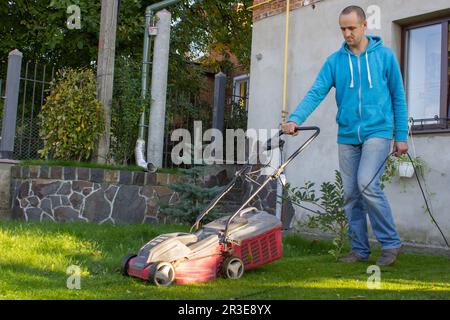 Ein Mann betreibt einen Rasenmäher im Hof in der Nähe des Hauses und mäht das Gras mit einem Rasenmäher Stockfoto
