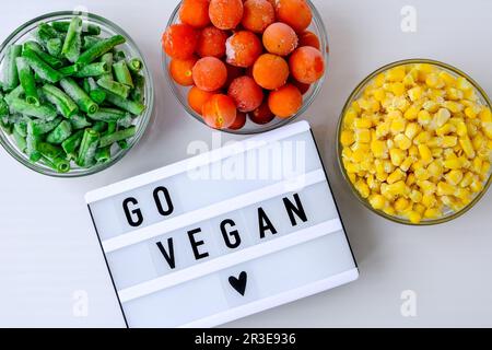 Lightbox mit Text GO GO VEGAN drei Schalen gefrorenes Gemüse Lebensmittel aus gelbem Mais, grünen Bohnen, roten Tomaten. Farben des Verkehrs l Stockfoto