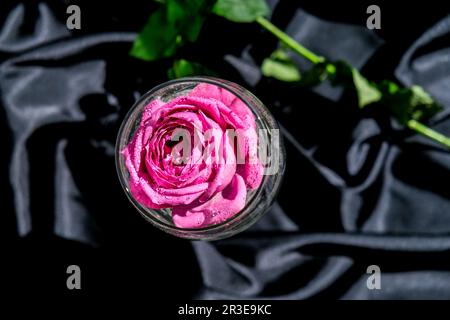 Weinglas gefüllt mit rosafarbenem Blumenpetalson-Tisch mit schwarzem Seidenstoff. Minimales, modernes Stillleben. Feiertagskonzept Valentinstag Stockfoto