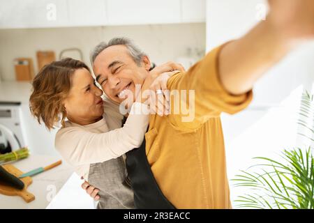 Fröhliches, altes Ehepaar, das Selfie macht, grimmig ist und sich in der Küche umarmt, Fotos von glücklichen Momenten macht Stockfoto