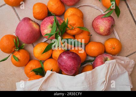 Rote Äpfel und Mandarinen in einem wiederverwendbaren Öko-Beutel auf Keramikboden. Obstlieferung vom Bauernmarkt, gesunde Lebensmittel. Blick von oben auf saftige Früchte. Stockfoto