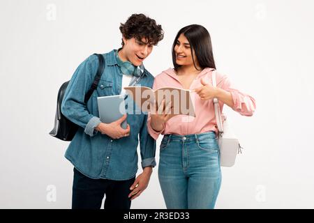 Fröhliche Schülerin, Die Dem Kerl Das Arbeitsbuch Über Weißen Hintergrund Zeigt Stockfoto