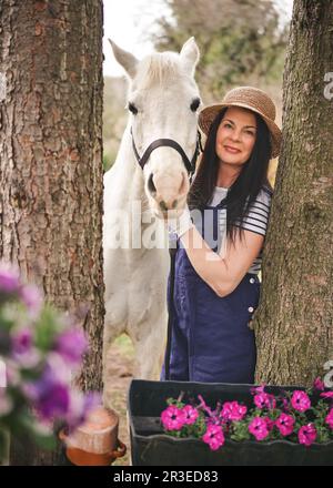 Frau in Arbeitskleidung und Strohhut, die neben ihrem weißen arabischen Pferd stand, Bäume an den Seiten, verschwommene Blumen im Vordergrund Stockfoto