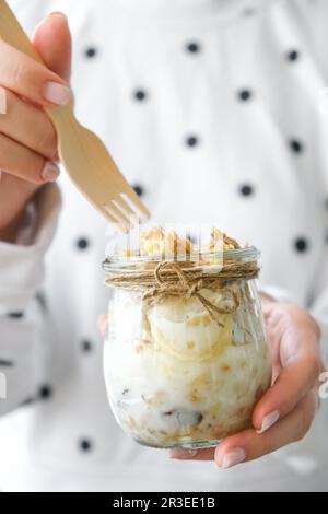 Weibliche Hände, Frau isst gesundes Frühstück. Haferflocken-Müsli mit griechischem Joghurt und Nüssen-Bananen-Müsli in Gläsern auf hellem Hintergrund Stockfoto