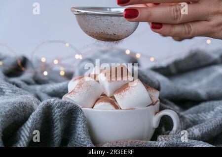 Hinzufügen von Kakaopulver auf heiße Schokolade mit Marshallows in weißer Tasse auf Pullover Hintergrund. Gemütliches weihnachtslicht. Stockfoto