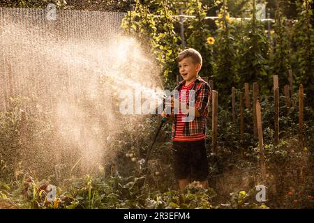 Der kleine Junge wäscht die Pflanzen und macht große Wasserspritzer Stockfoto