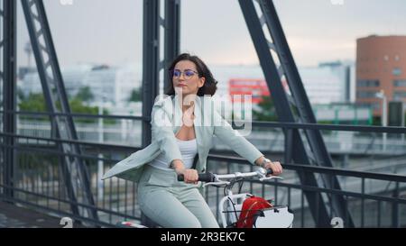 Junge, lockere und glückliche Geschäftsfrau fährt morgens gerne Fahrrad in der Stadt auf einer Brücke mit einer wunderschönen Stadtlandschaft Stockfoto