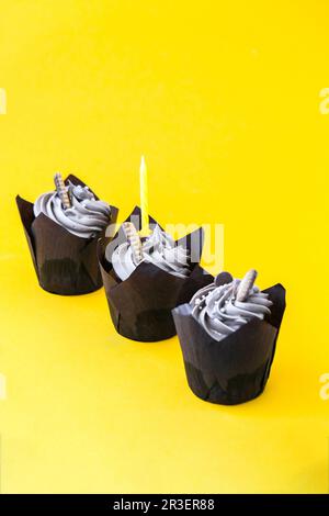 Leckere Cupcakes aus Schokolade mit Creme und Kerze auf gelbem Hintergrund. Drei Schokoladenmuffin. Geburtstagsparty. Stockfoto