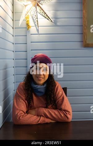 Junge dunkelhaarige Frau in einem orangefarbenen Pullover mit Glitzer, trägt eine mehrfarbige Mütze mit blauem Schal, sitzt in einem Haus, mit einem Stern Stockfoto