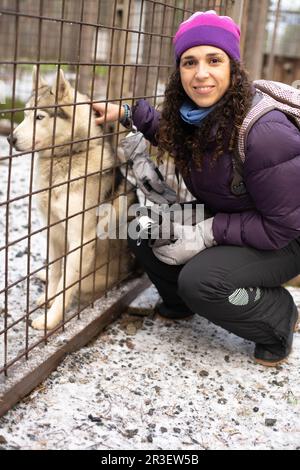 Junge brünette Frau kniet in Winterkleidung und hat einen Husky-Hund hinter einem Zaun gestreichelt Stockfoto