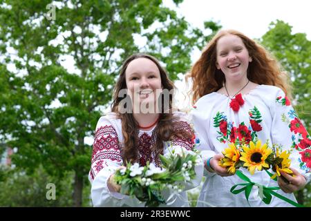 Wunderschöne junge Mädchen, die bestickte Hemden tragen und lachen, Kränze auf die wassergerichtete Kamera legen, die Kränze auf uns strecken, verschiedene Sonnenblumen, Feldblumen, Mädchen hat rotes, helles, sonniges Haar Stockfoto