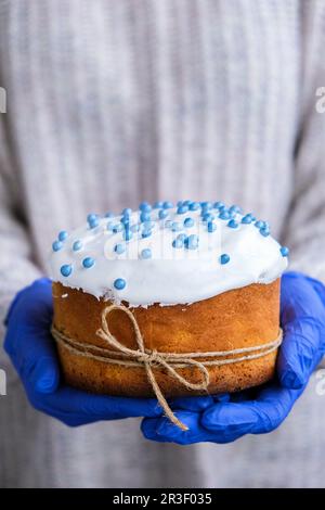 Hände in blauen Handschuhen halten Osterkuchen mit weißem Belag und blauen Streuseln. Frau mit traditionellem russischen Osterkuchen. Nach Hause Stockfoto