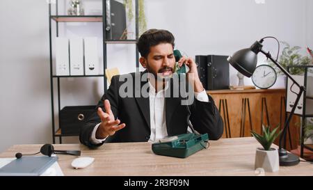 Ein bärtiger indischer Geschäftsmann, der mit einem Kunden über ein altmodisches Kabeltelefon am Arbeitsplatz des Heimbüros sprach. Verkäufer im formellen Anzug. Entfernter Job. Erwerbstätigkeit Stockfoto