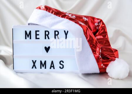 Lightbox mit Text FROHE WEIHNACHTEN mit weihnachtsmütze auf Seidenstoff-Hintergrund. Konzept für Winterurlaub. Stockfoto