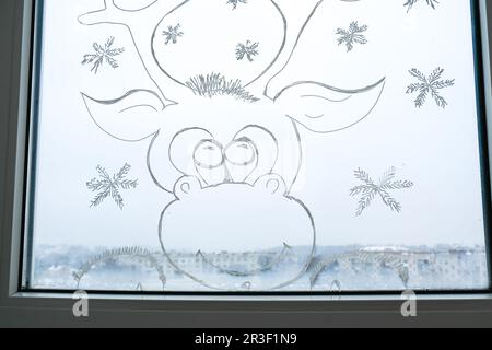 Weihnachtshirsche auf Fensterglas gemalt. Weihnachtsdekor im Winter auf dem Fensterglas. Weißes Muster von Hirschen. Schneekunst-Tracery-Wind Stockfoto