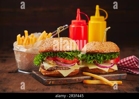 Zwei hausgemachte Burger mit Rind-, Käse- und Zwiebelmarmelade auf einem Holzbrett, Pommes in einem Metallkorb und Saucen. Fast Food Konz Stockfoto