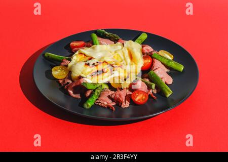 Köstlicher Raclette-Käse, serviert über Rinderbraten mit Tomaten und Spargel, Gericht auf rotem Hintergrund. Hartes Licht, tiefer Schatten Stockfoto