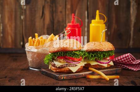 Zwei hausgemachte Burger mit Rind-, Käse- und Zwiebelmarmelade auf einem Holzbrett, Pommes in einem Metallkorb und Saucen. Fast Food Konz Stockfoto