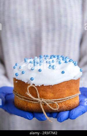 Hände in blauen Handschuhen halten Osterkuchen mit weißem Belag und blauen Streuseln. Frau mit traditionellem russischen Osterkuchen. Nach Hause Stockfoto