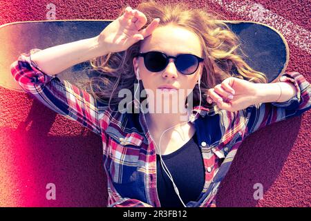 Millennials-Hippie-Frau mit Sonnenbrille und Kopfhörern, die auf einem Skateboard auf der Straße liegt. Skater-Mädchen auf einem Longboard. Cool fe Stockfoto