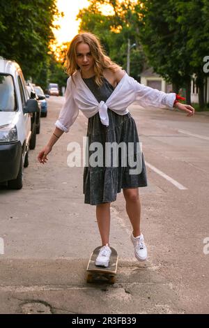 Millennials Frau in Kleidern, die auf einem Skateboard auf der Straße fährt. Skater-Mädchen auf einem Longboard. Coole Skateboarderin bei Sonnenuntergang. Carefre Stockfoto