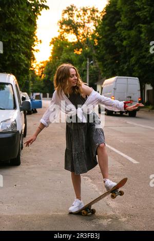 Millennials Frau in Kleidern, die auf einem Skateboard auf der Straße fährt. Skater-Mädchen auf einem Longboard. Coole Skateboarderin bei Sonnenuntergang. Carefre Stockfoto