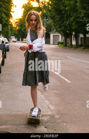 Millennials Frau in Kleidern, die auf einem Skateboard auf der Straße fährt. Skater-Mädchen auf einem Longboard. Coole Skateboarderin bei Sonnenuntergang. Carefre Stockfoto