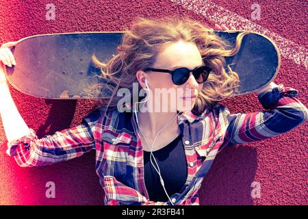 Millennials-Hippie-Frau mit Sonnenbrille und Kopfhörern, die auf einem Skateboard auf der Straße liegt. Skater-Mädchen auf einem Longboard. Cool fe Stockfoto