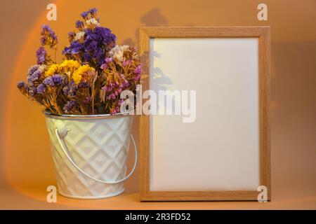 Holzrahmen mit leerem weißem Papier und zarten Trockenblumen in weißem Eimer auf beigefarbenem Hintergrund. Minimaler trendiger CO Stockfoto