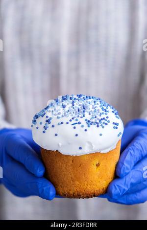 Hände in blauen Handschuhen halten Osterkuchen mit weißem Belag und blauen Streuseln. Frau mit traditionellem russischen Osterkuchen. Nach Hause Stockfoto