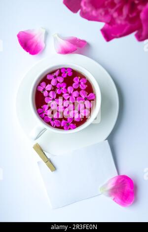 Kreatives Layout mit pinkfarbenen Pfingstrosen und einer Tasse Tee auf einem hellen Tisch. Eine Notiz aus Papier. Speicherplatz kopieren. Frühling, saisonaler valentinstag, wom Stockfoto