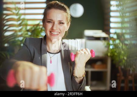 Nachhaltiger Arbeitsplatz. Glückliche, moderne Eigentümerin eines kleinen Unternehmens in einem modernen grünen Büro mit Kurzhanteln. Stockfoto