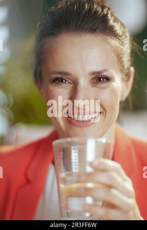 Nachhaltiger Arbeitsplatz. Glückliche, moderne Eigentümerin eines kleinen Unternehmens in einem modernen grünen Büro in einer roten Jacke mit einer Tasse Wasser. Stockfoto