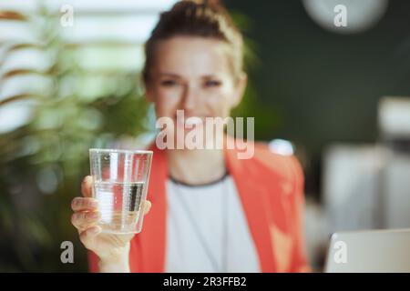 Nachhaltiger Arbeitsplatz. Lächelnde, moderne Eigentümerin eines kleinen Unternehmens bei der Arbeit in einer roten Jacke mit einer Tasse Wasser. Stockfoto