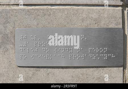 Silberplatte mit Braille-Text an grauer Wand, Nahaufnahme Stockfoto