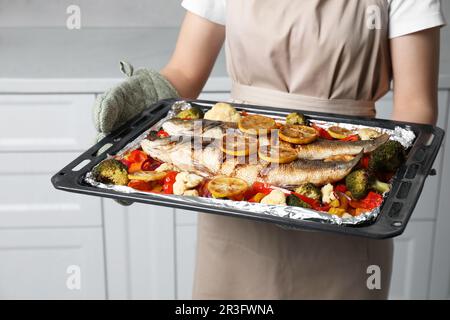 Eine Frau, die ein Backblech mit Seebarsch-Fisch und Gemüse in der Küche hält, Nahaufnahme Stockfoto