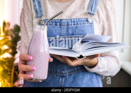 Weibliche Hände mit Blueberry Smoothie garniert mit Blaubeeren. Eine Frau, die ein Glas Frühstücksprotein-Smoothie isst und liest Stockfoto