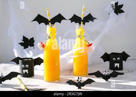 Halloween-Mocktails. Oranger alkoholfreier Cocktail für Halloween-Party auf weißem Tisch. Die Trinkhalme sind mit geschnittenen Papierfledermäusen verziert. Stockfoto