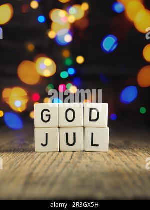 Kleine Holzklötze mit der Aufschrift Frohe Weihnachten auf skandinavisch. Verschwommenes Weihnachtslicht im Hintergrund, saisonaler Hintergrund für Feiertage Stockfoto