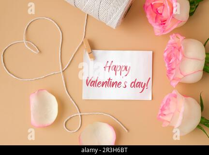 Zarte rosa Rosen mit Spule aus weißem Baumwollseil in Herzform auf beigefarbenem Hintergrund. Papiernotiz mit Text ALLES GUTE ZUM VALENTINSTAG. Stockfoto