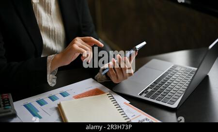 Nahaufnahme einer Geschäftsfrau, die auf ihrem Telefon scrollt, E-Mails oder Nachrichten auf ihrem Telefon abruft, während sie an ihrem Schreibtisch sitzt. Stockfoto