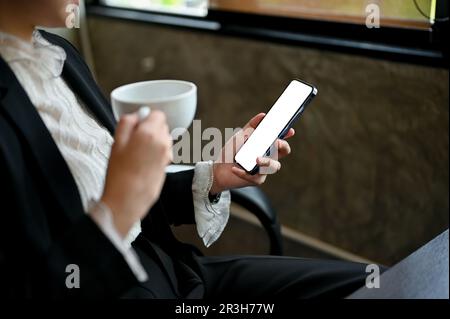 Nahaufnahme einer professionellen Geschäftsfrau, die ihren Morgenkaffee trinkt, während sie E-Mails auf ihrem Telefon an ihrem Schreibtisch im Büro abruft. Smartphone Stockfoto