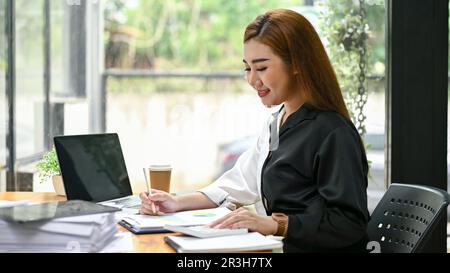 Seitenansicht einer schönen und lächelnden asiatischen Millennials-Geschäftsfrau, die Geschäftsberichte an ihrem Schreibtisch im Büro prüft. Stockfoto