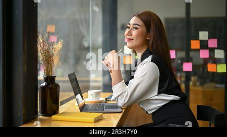 Seitenansicht einer glücklichen und lächelnden asiatischen Geschäftsfrau der Millennials, die aus dem Fenster schaut und von ihrem Glück träumt, während sie aus der Ferne in einem Coff arbeitet Stockfoto