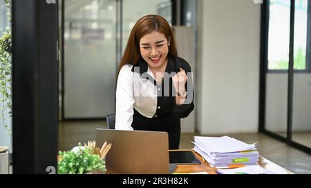 Fröhliche und aufgeregte asiatische Geschäftsfrau der Millennials zeigt geballte Faust, während sie auf ihren Laptop-Bildschirm am Schreibtisch blickt und ihr Projekt als Erfolg feiert Stockfoto