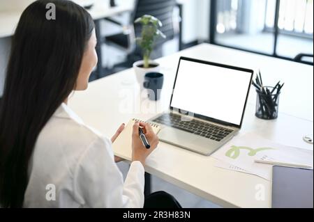 Nahaufnahme Rückansicht, Eine professionelle und fokussierte asiatische Geschäftsfrau oder Büroangestellte, die sich auf ihrem Notebook Notizen macht, während sie auf ihren Schoß blickt Stockfoto