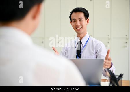 Glücklicher und lächelnder asiatischer Geschäftsmann, der seinem Kollegen Daumen hoch zeigt, während er im Büro zusammen arbeitet. Stockfoto