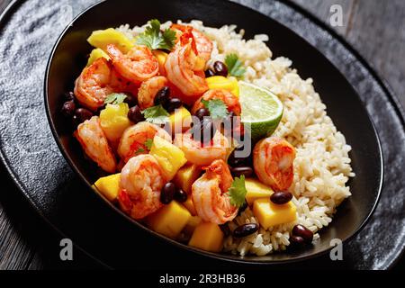 Karibische gegrillte Garnelen mit schwarzen Bohnen, Mango-Ananassalsa serviert mit braunem Reis in schwarzer Schüssel, Nahaufnahme Stockfoto